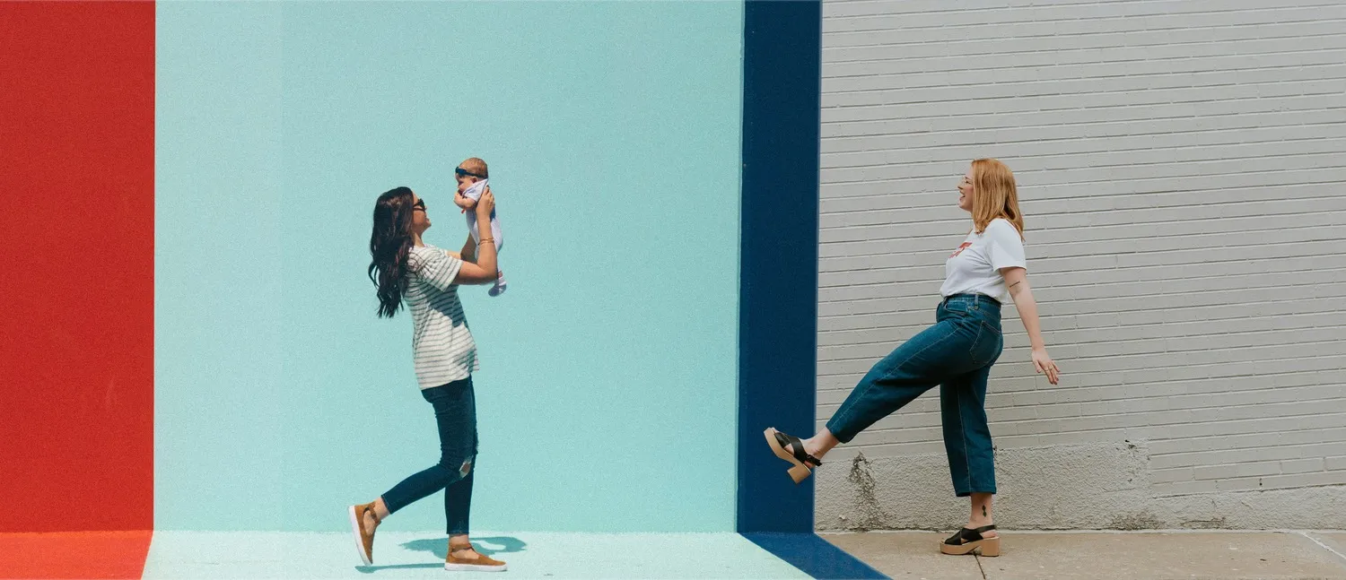 Two mothers outdoors against a painted wall, one lifting her baby overhead