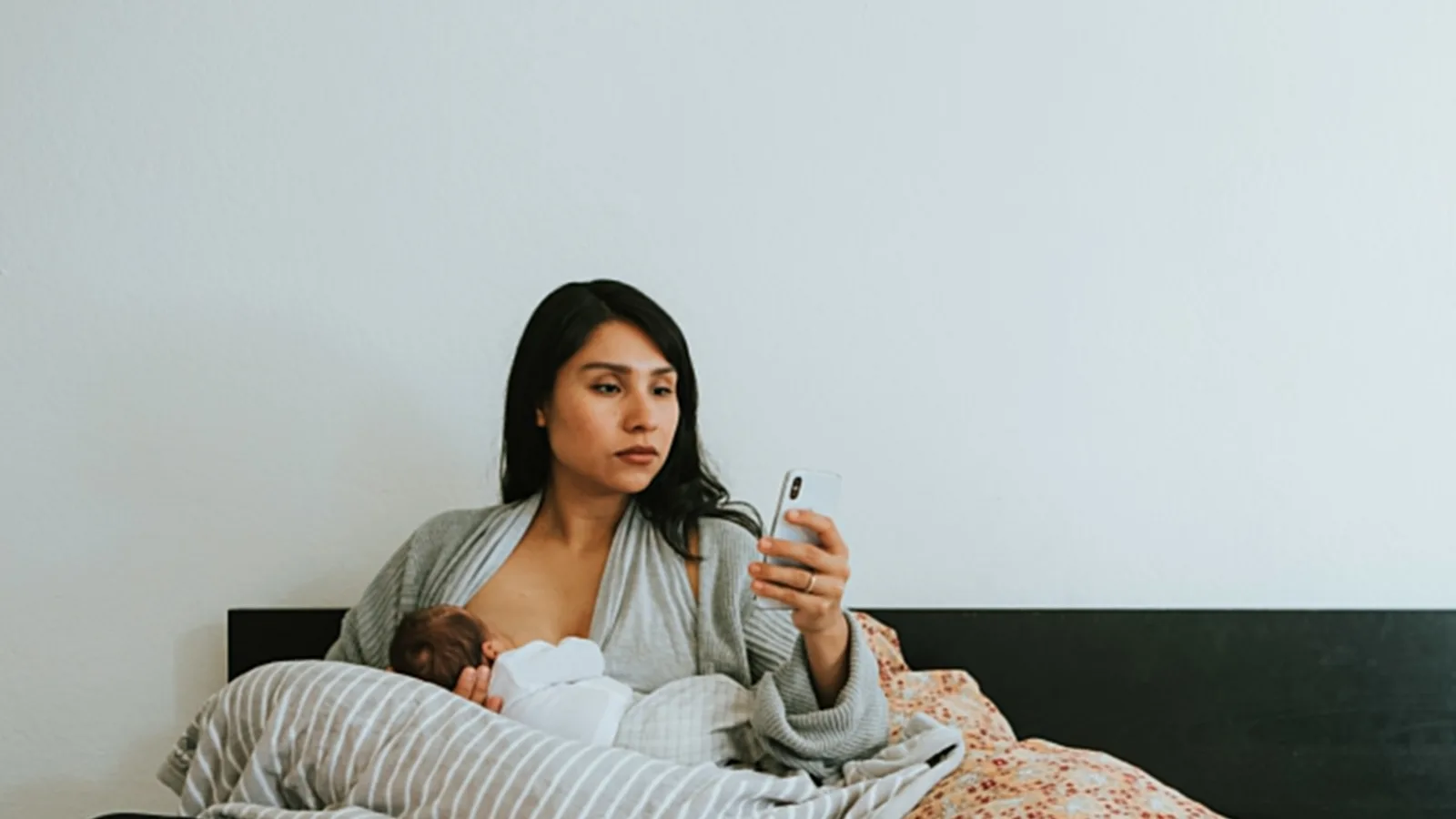A mother nursing her baby in bed while scrolling on her phone